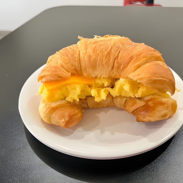 A golden croissant breakfast sandwich with scrambled eggs and melted cheese on a white plate.