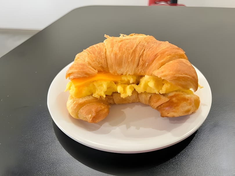 A golden croissant breakfast sandwich with scrambled eggs and melted cheese on a white plate.