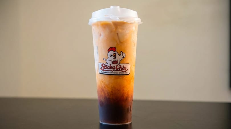 Thai iced tea in a clear plastic cup with the "Sticky Chix" logo on a dark surface.