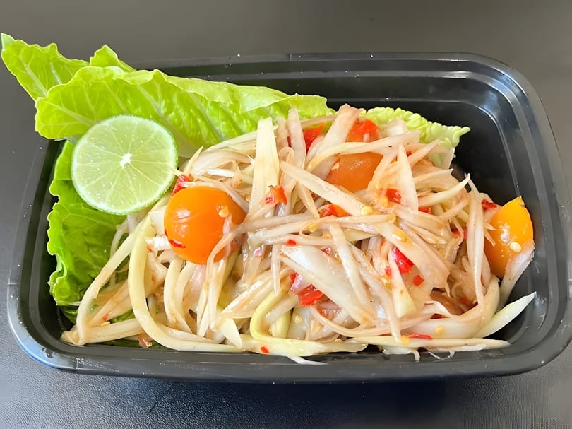 Som Tum (papaya salad) with lime, cherry tomatoes, and lettuce in a black container.