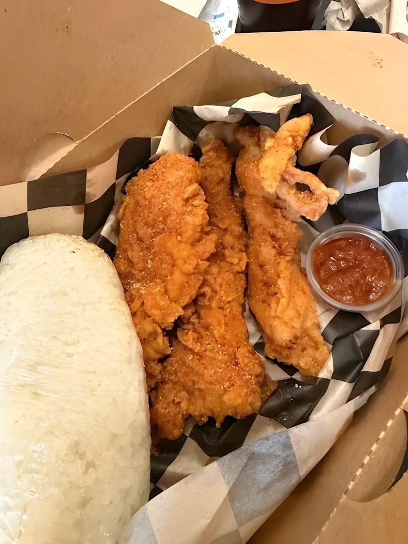Fried chicken tenders, white rice, and a side of sauce in a takeout box with checkered paper.