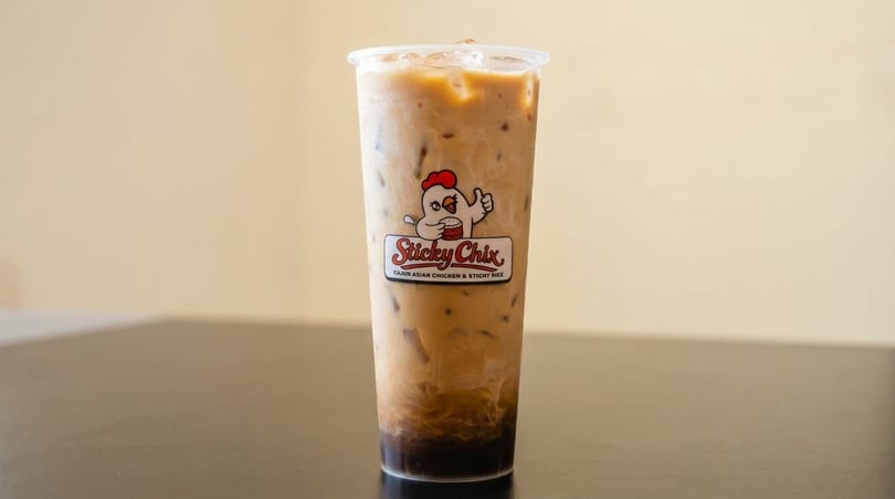 Iced coffee in a clear plastic cup with the "Sticky Chix" logo on a dark table.