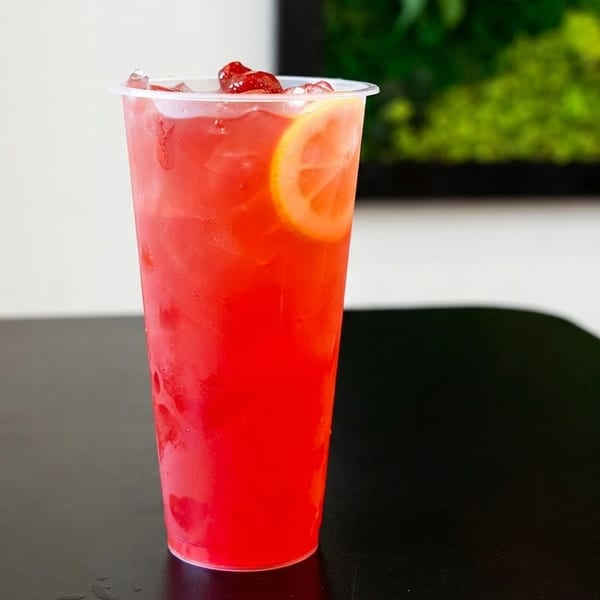 A vibrant red drink in a clear plastic cup, garnished with a lemon slice and berries.