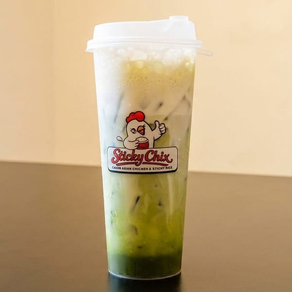 Iced green drink with white foam and the Sticky Chix logo on a dark table.