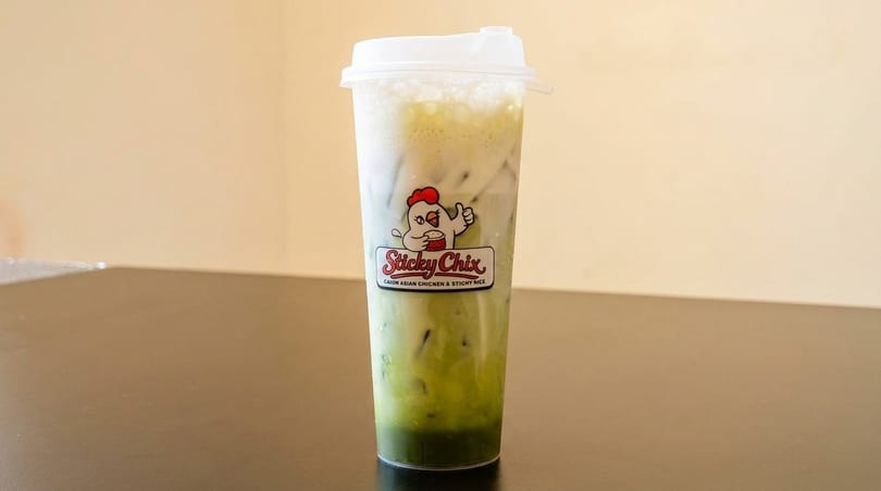 Iced green drink with white foam and the Sticky Chix logo on a dark table.