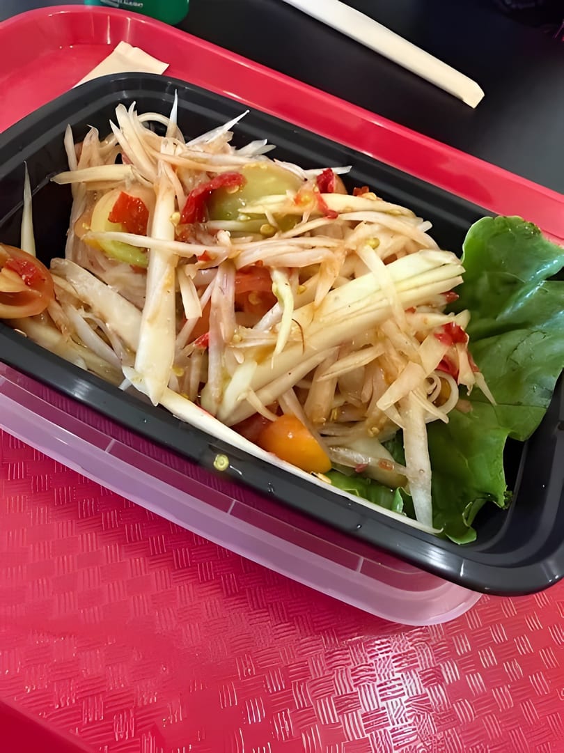 A close-up of a vibrant papaya salad with tomatoes and lettuce in a black to-go container.