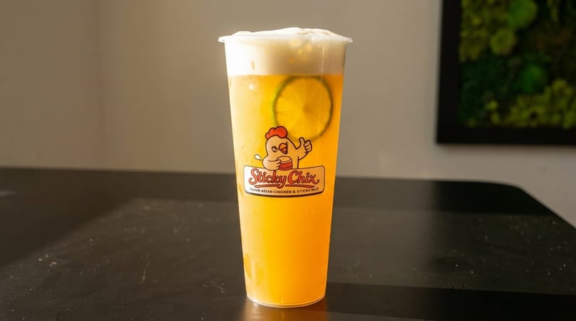 A tall plastic cup of yellow iced tea with a lime slice and foam, featuring the Sticky Chix logo.
